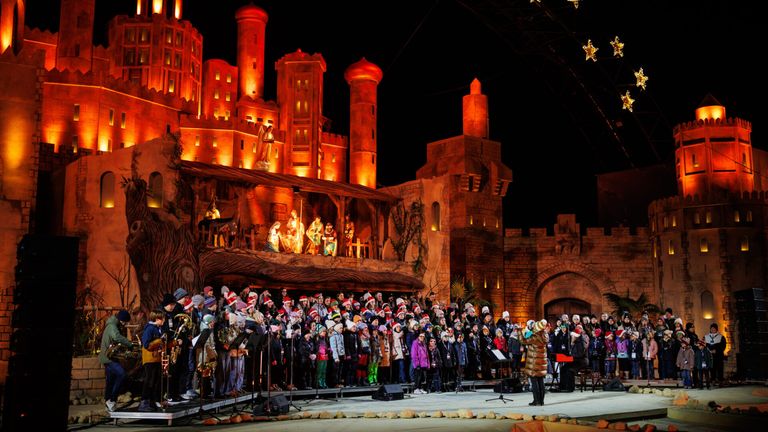 Ein Chor, gekleidet in Wintermützen und Mänteln, tritt auf einer Bühne vor einer beleuchteten Burg bei Nacht auf.