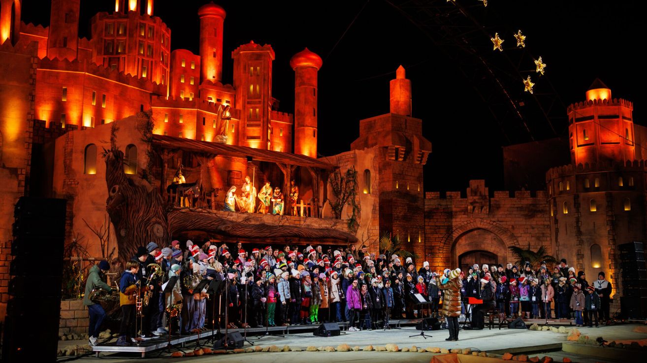 Ein Chor, gekleidet in Wintermützen und Mänteln, tritt auf einer Bühne vor einer beleuchteten Burg bei Nacht auf.