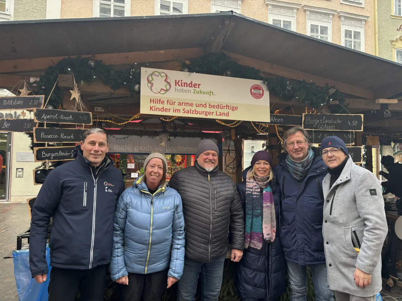 Five people are standing in front of a sign that says Hilfe fur arm und bedruftige Kinder im Salzburger Land. They are all wearing jackets and some hats. They seem to be posing for a photo. Behind them is a building with a Christmas decoration.