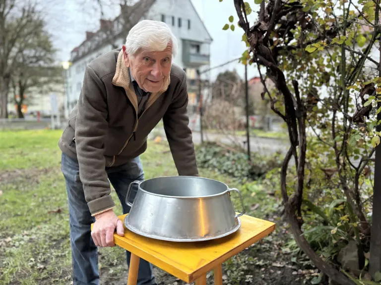 Ein älterer Mann steht im Freien in einem Garten und betrachtet eine Edelstahlschüssel auf einem gelben Tisch.