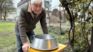 An older man is outdoors, standing in a garden, and examining a stainless steel bowl on a yellow table.