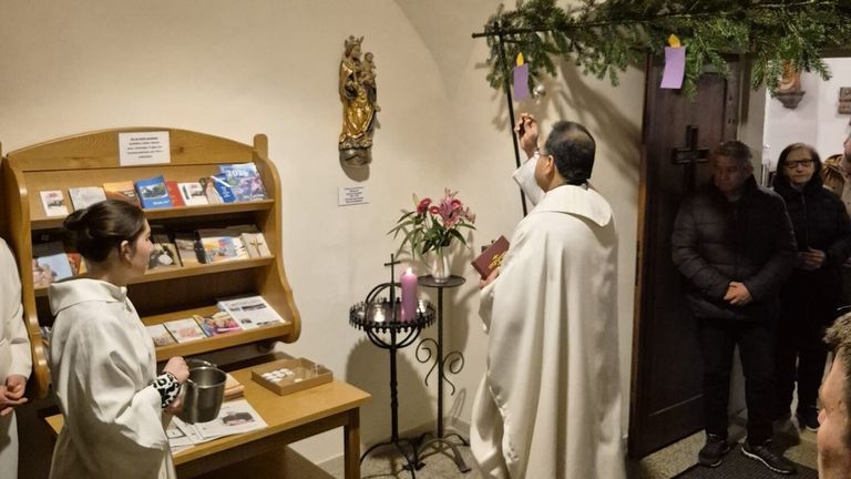 Ein Priester zündet eine Kerze in einer Kapelle an. Der Raum ist mit religiösen Artefakten dekoriert. Ein Tisch hält ein Buch und eine Blumenvase.