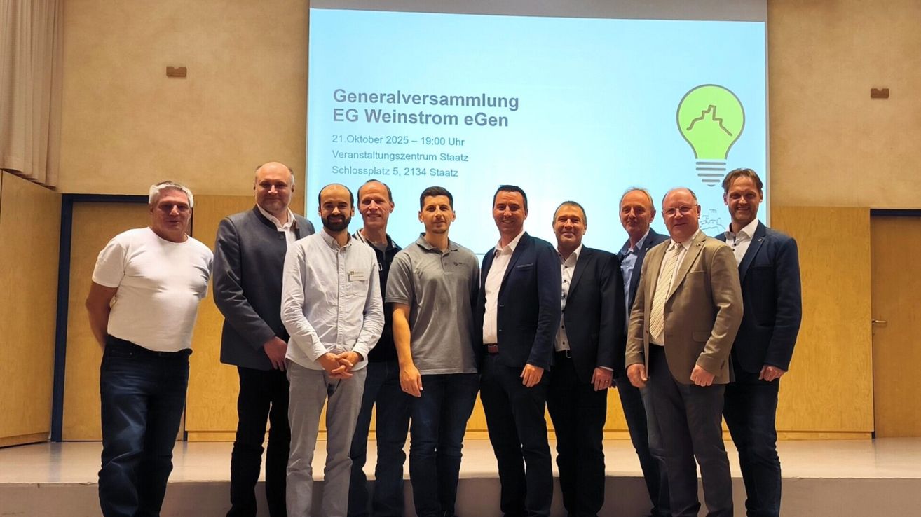 Sieben Männer stehen auf einer Bühne vor einer Leinwand mit einer Präsentation über eine Generalversammlung von EG Weinstrom eGen.