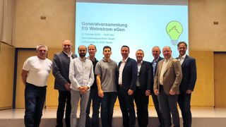 Seven men stand on a stage in front of a screen with a presentation about a general meeting of EG Weinstrom eGen.