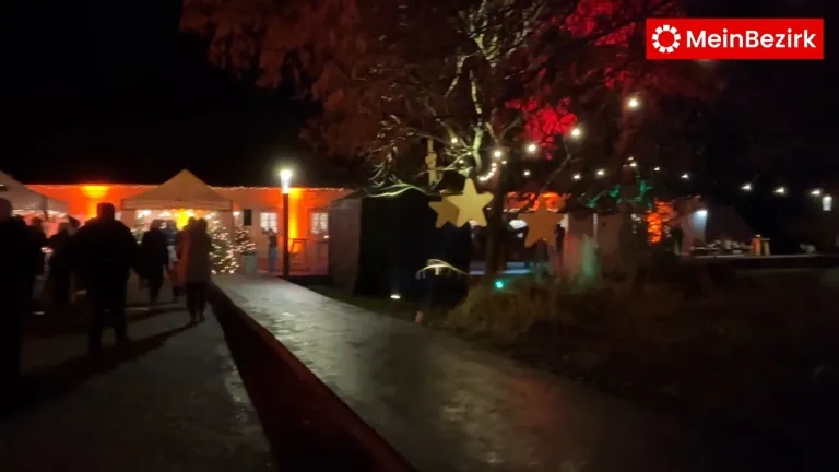 Bei Nacht ist ein Gehweg von Lichtern beleuchtet. Eine Person geht auf dem Bürgersteig. Bäume sind mit Lichtern geschmückt, und es gibt ein Zelt mit einem Stern. Ein Gebäude ist im Hintergrund sichtbar.