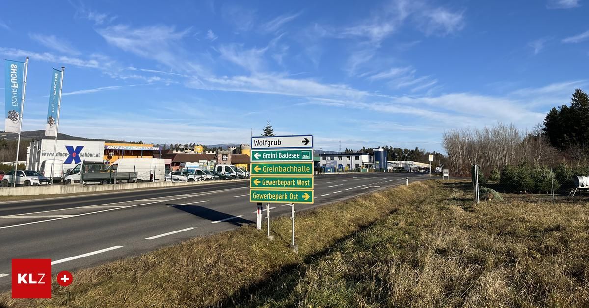 Ein Straßenschild am Straßenrand zeigt Richtungen nach Wolfgrub, Grein Badsee, Greinbachhalle und Gewerbepark West. Im Hintergrund sind geparkte Autos und Gebäude zu sehen.