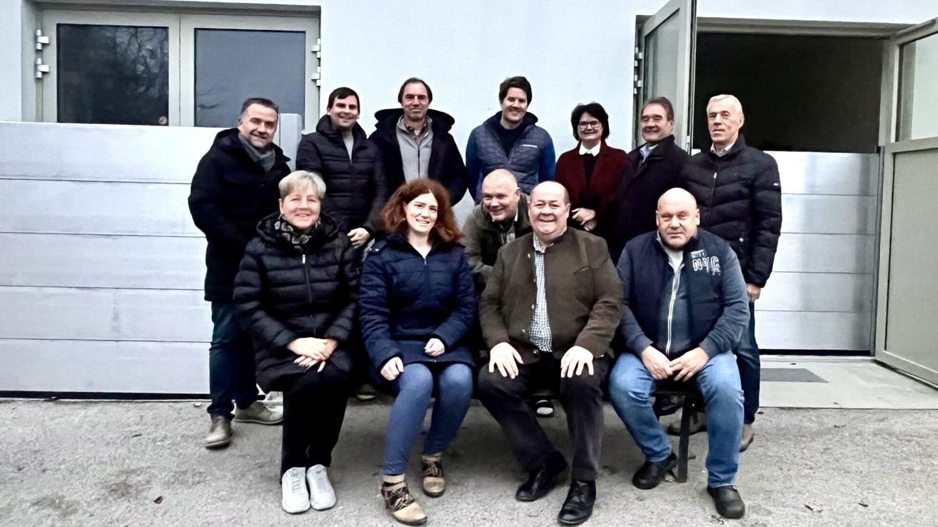 A group of people dressed warmly for cold weather, posing for a photo in front of a building with an open door. They are sitting and standing in two rows.
