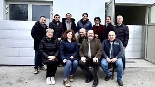 A group of people dressed warmly for cold weather, posing for a photo in front of a building with an open door. They are sitting and standing in two rows.