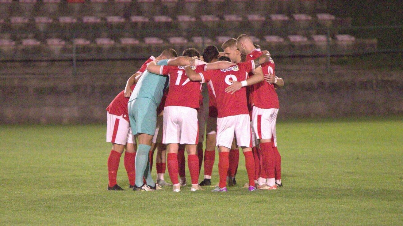 A soccer team huddles together on a field, wearing red uniforms, with one player in a light blue jersey.