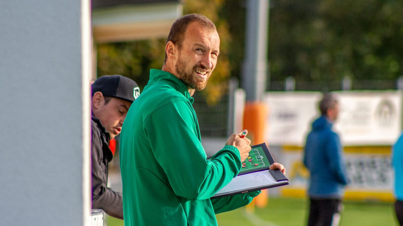 Ein Fußballtrainer lächelt und hält eine Mappe mit einer Zeichnung eines Fußballfeldes. Ein anderer Mann sieht ihn an.