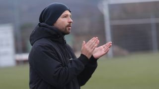 A man in a black jacket and beanie is clapping on a field. The background is blurred.