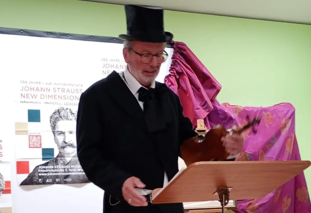 An older man in formal attire, including a top hat and bow tie, is playing a violin at a podium. A backdrop displays an image of Johann Strauss.