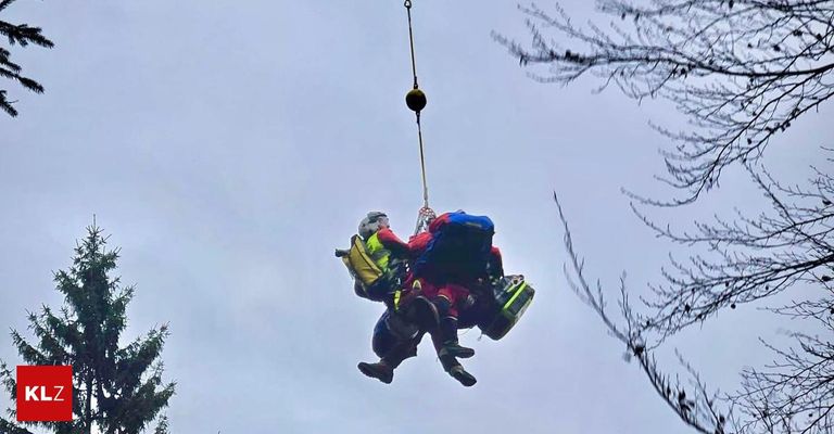 Zwei Personen werden von einem Hubschrauber vom Boden gehoben. Beide tragen Helme und haben Rucksäcke dabei.