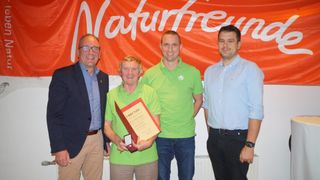 Four men in green and blue shirts stand in front of a banner. The man in the center holds a certificate and an award. The man on the left wears a suit and glasses.
