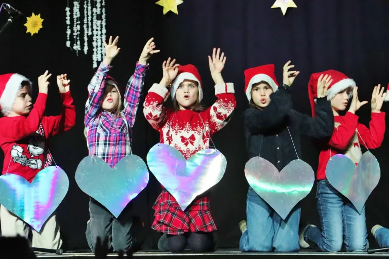 Fünf Kinder in festlicher Kleidung knien auf einer Bühne, halten herzförmige Schilder und heben die Hände, möglicherweise bei einem Weihnachtsevent auftretend.