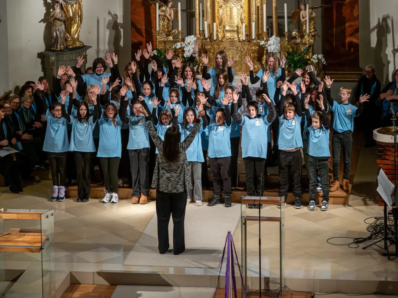 A choir of children dressed in blue shirts is performing on stage. A woman stands before them, raising her hands. Behind the choir, a golden altar with candles is visible.