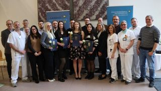 A group of individuals, likely medical students, are posing for a photo. They hold certificates and flowers, standing in front of a backdrop with logos.