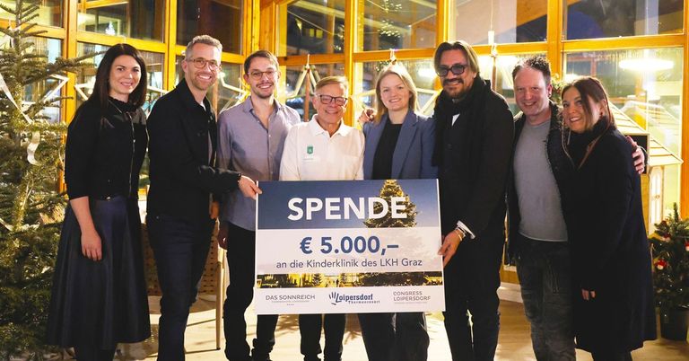Sechs Personen lächeln für ein Foto und halten ein Schild mit der Aufschrift 'Spende € 5.000,' für die Kinderklinik des LKH Graz. Sie stehen in einem Raum mit gelben Glasfenstern.