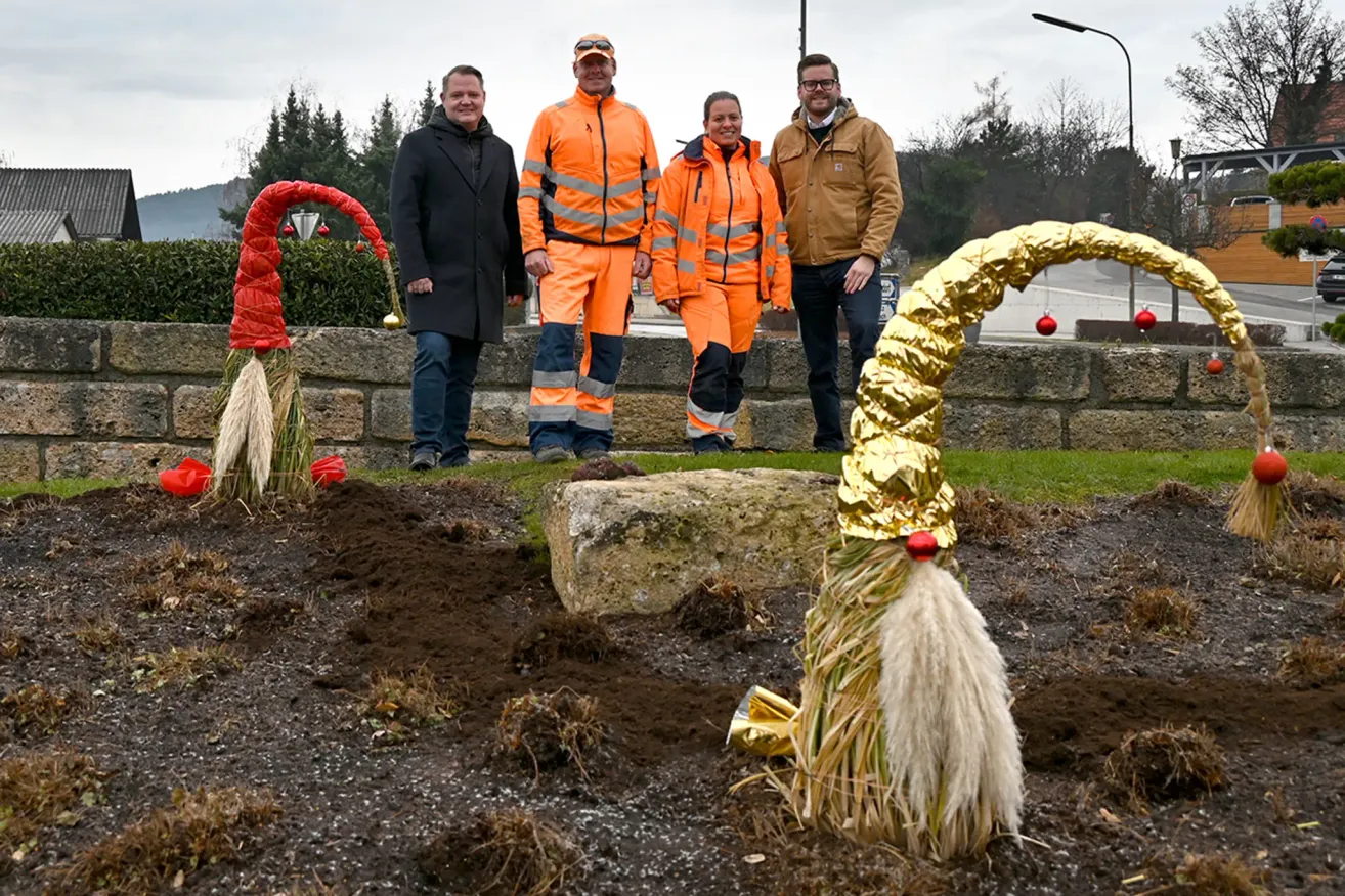 Five people stand outdoors on a stone wall, wearing high-visibility clothing. One wears a reflective orange jumpsuit, another a black coat. Behind them, a golden sculpture and decorative elements are visible. Trees and a cloudy sky form the background.