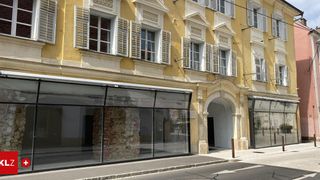 A yellow building with white shutters has a brick wall in the middle of the glass entrance. It has a stone archway entrance.