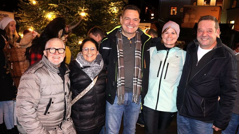 Vier Personen stehen lächelnd vor einem Weihnachtsbaum. Der Mann in der Mitte trägt einen Schal, und die Frau rechts trägt eine Mütze. Im Hintergrund befindet sich ein Gebäude mit einem Fenster und einer Straßenlaterne.