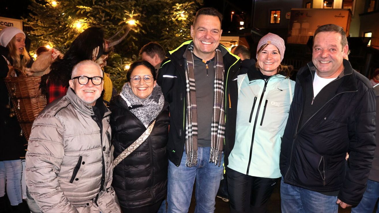 Vier Personen stehen lächelnd vor einem Weihnachtsbaum. Der Mann in der Mitte trägt einen Schal, und die Frau rechts trägt eine Mütze. Im Hintergrund befindet sich ein Gebäude mit einem Fenster und einer Straßenlaterne.