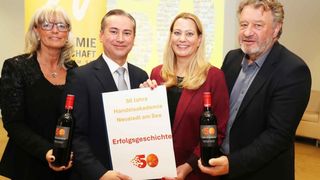 Vier Personen in formeller Kleidung halten ein Schild, das 50 Jahre der Handelsakademie Neusiedl am See feiert, zwei Männer halten Weinflaschen.