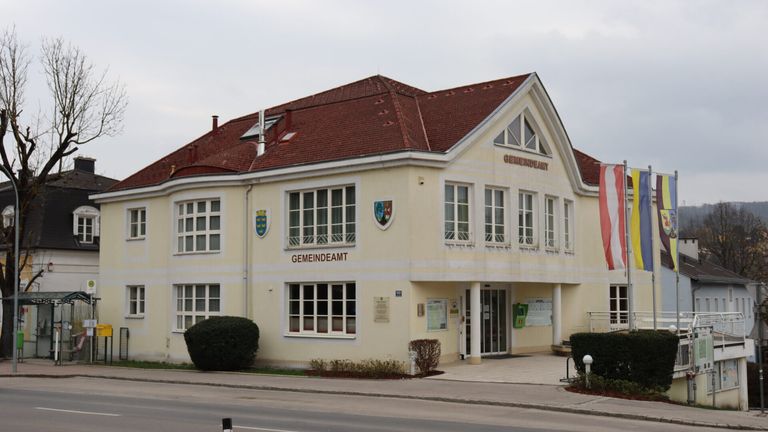 Ein Gemeindeamtsgebäude mit rotem Dach und weißen Wänden. Es hat viele Fenster und zwei Wappen über dem Eingang.