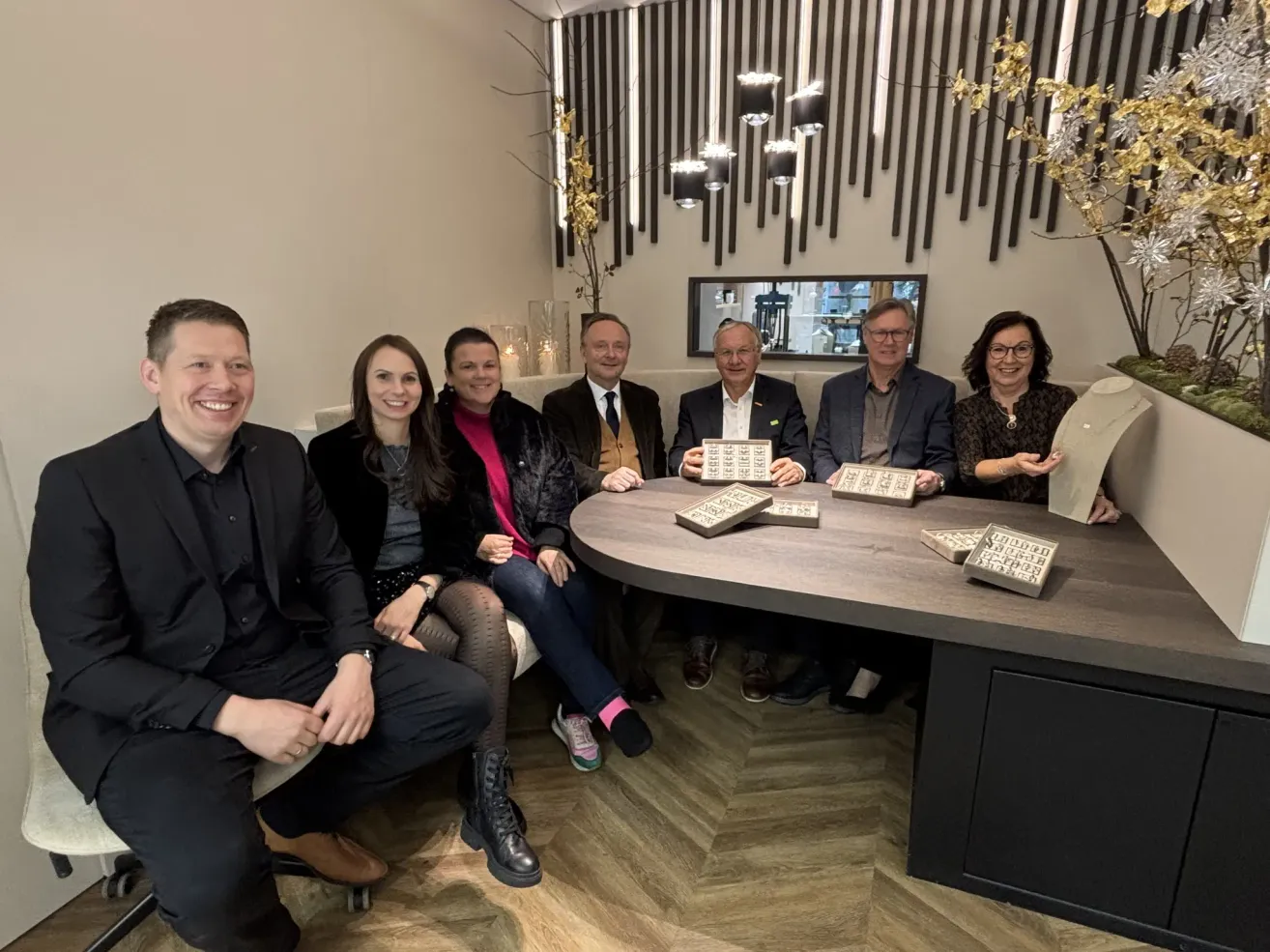 A group of people pose for a photo inside a modern room. They are seated on a bench around a table, with puzzles on it. The room has wooden flooring and decorative elements on the wall.