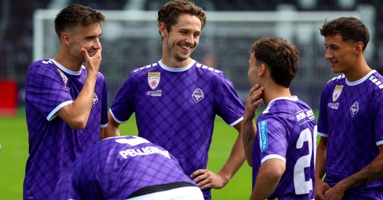 Eine Gruppe männlicher Fußballspieler in lila Trikots steht auf einem Feld, lächelt und lacht, wobei ein Spieler ein weißes Shirt mit Logo trägt.