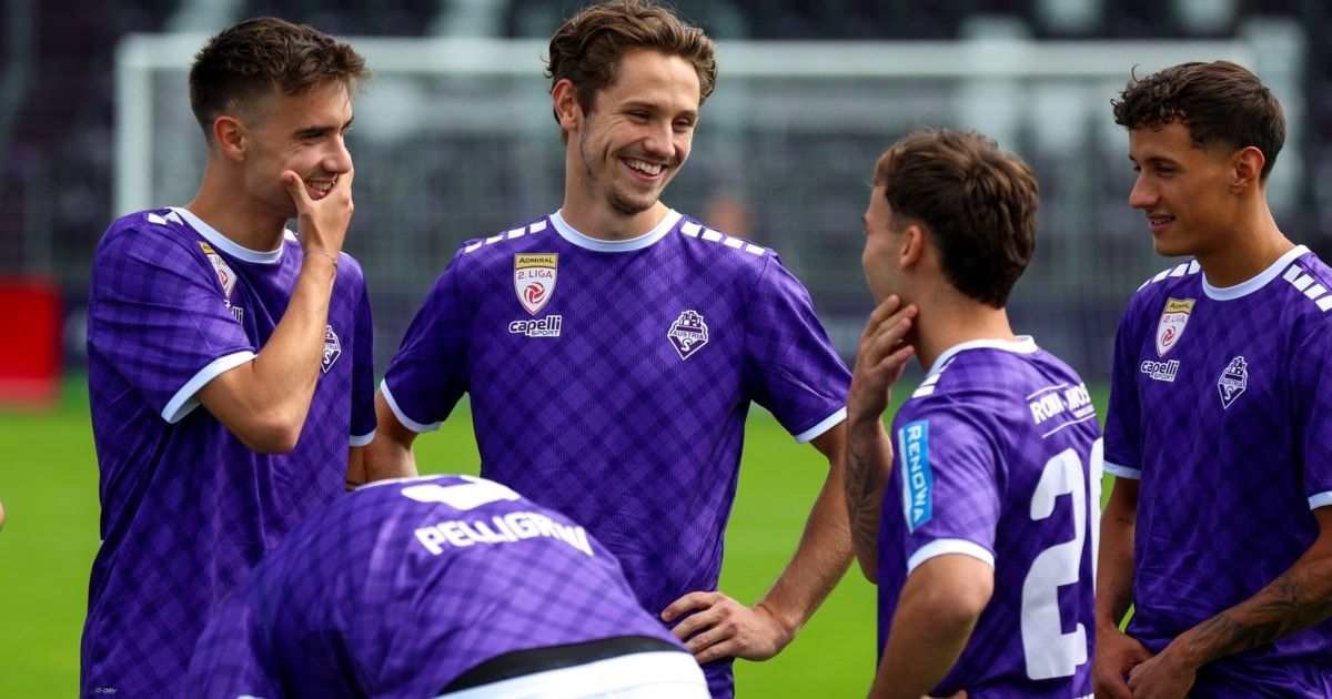 Eine Gruppe männlicher Fußballspieler in lila Trikots steht auf einem Feld, lächelt und lacht, wobei ein Spieler ein weißes Shirt mit Logo trägt.