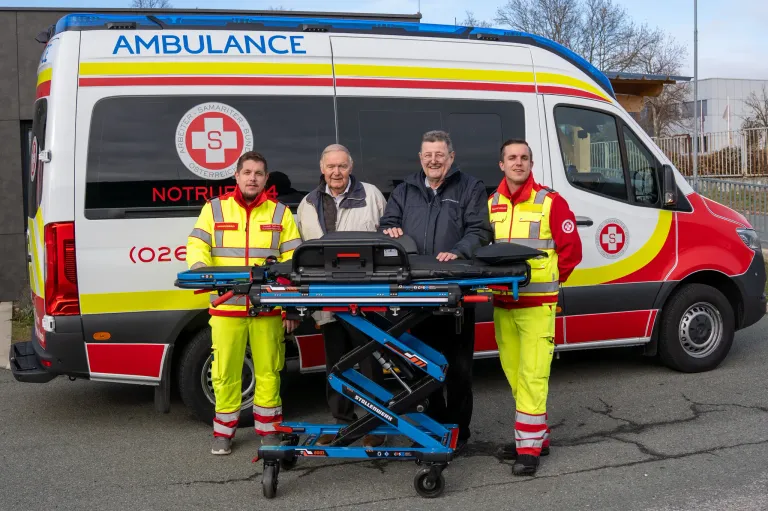 Drei Männer in gelben und roten Uniformen stehen neben einem blauen Wagen mit einem Krankenwagen im Hintergrund. Der Krankenwagen hat ein rot-weißes Design mit einem Logo, das einen 'S' und 'Oesterreich' Text zeigt.