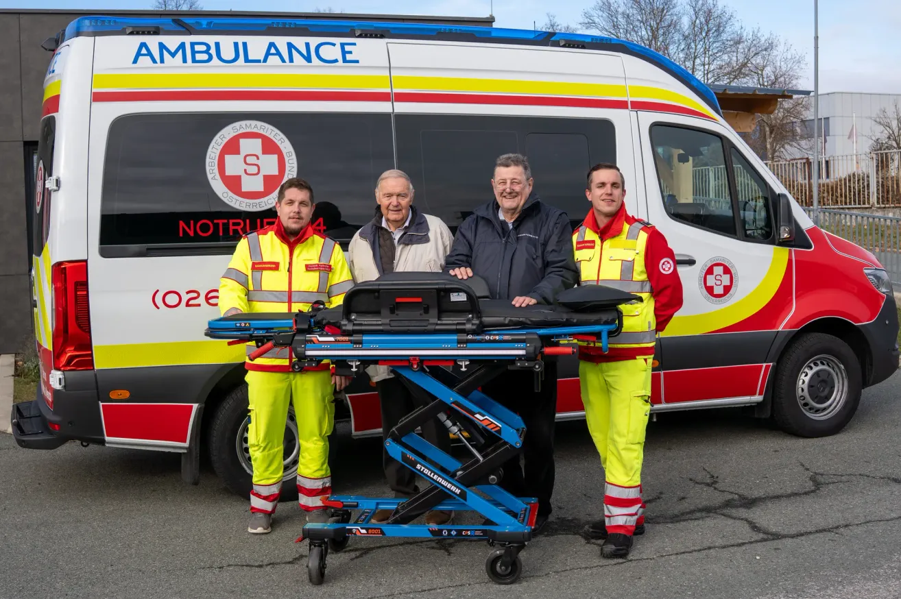 Drei Männer in gelben und roten Uniformen stehen neben einem blauen Wagen mit einem Krankenwagen im Hintergrund. Der Krankenwagen hat ein rot-weißes Design mit einem Logo, das einen 'S' und 'Oesterreich' Text zeigt.