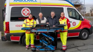 Drei Männer in gelben und roten Uniformen stehen neben einem blauen Wagen mit einem Krankenwagen im Hintergrund. Der Krankenwagen hat ein rot-weißes Design mit einem Logo, das einen 'S' und 'Oesterreich' Text zeigt.