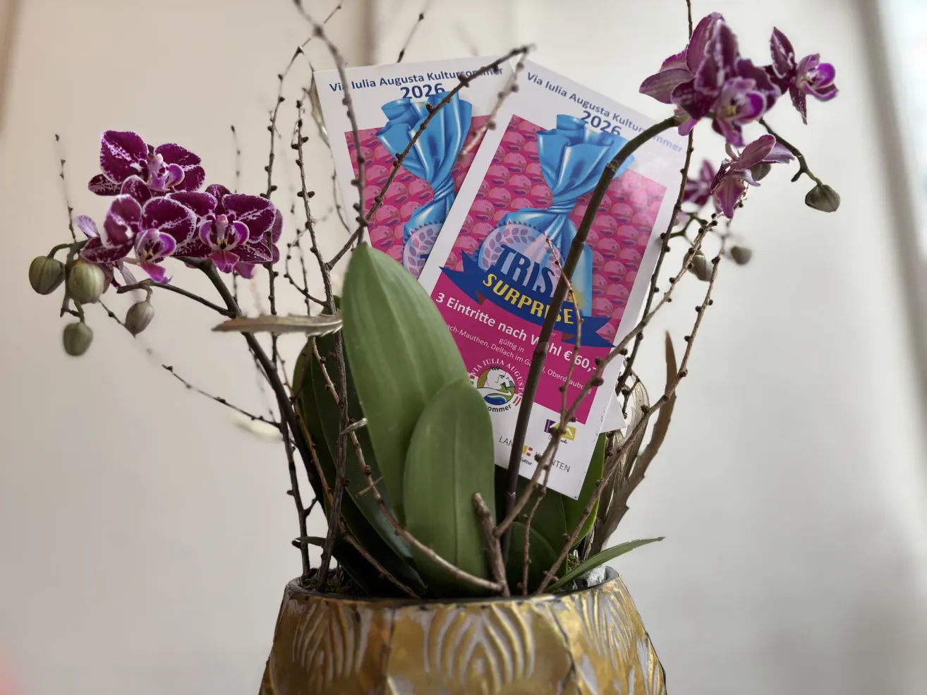 In einer goldenen Vase befindet sich ein Strauß aus lila Orchideen und grünen Blättern. Zwei Tickets für 'Julia Augusta Kultursommer 2026' sind an den Orchideenstielen befestigt. Die Tickets haben ein rosa-blaues Design mit dem Text 'TRIS SURPRISES'.
