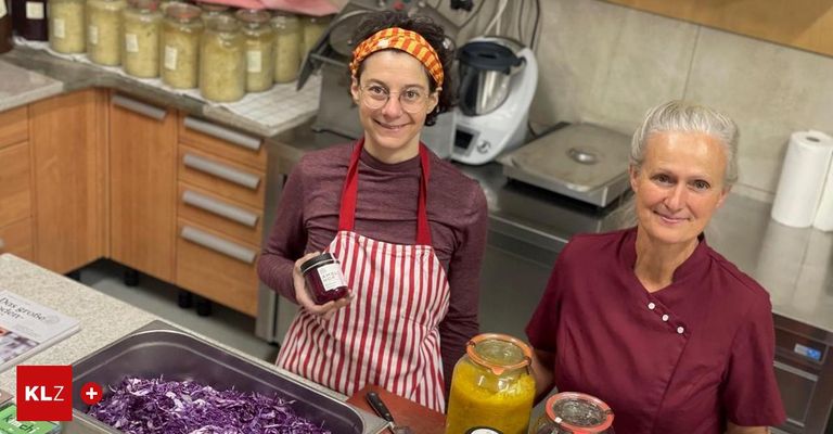Zwei Frauen in einer Küche, eine hält eine Dose und lächelt. Hinter ihnen stehen Gläser und Küchengeräte. Ein Tablett mit rotem Kohl auf der linken Seite und zwei Glasgläser mit Deckeln auf der rechten Seite.