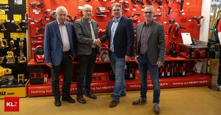 Vier Männer in formeller Kleidung stehen vor einer roten Wand mit Werkzeug und Ausrüstung. Zwei Männer schütteln sich die Hände, während die anderen neben ihnen stehen und lächeln.