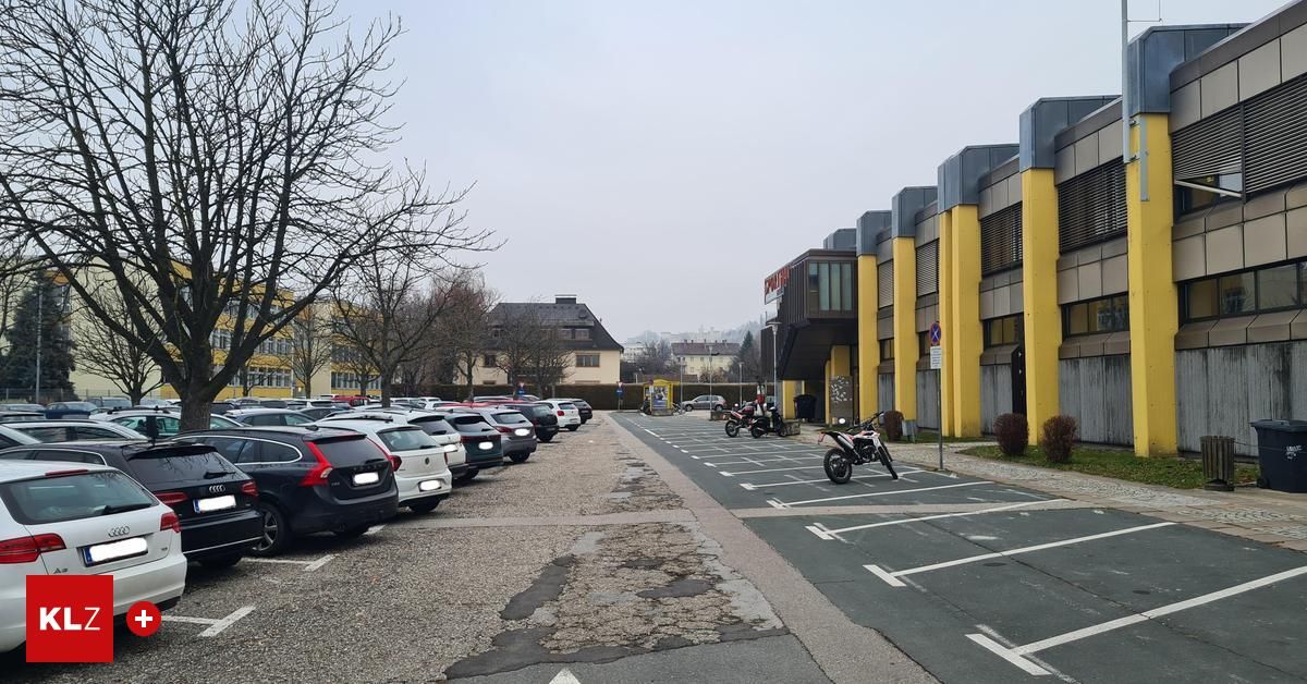 An empty parking lot with a row of parked cars, two motorcycles parked at the end, a yellow building, and a small bush.