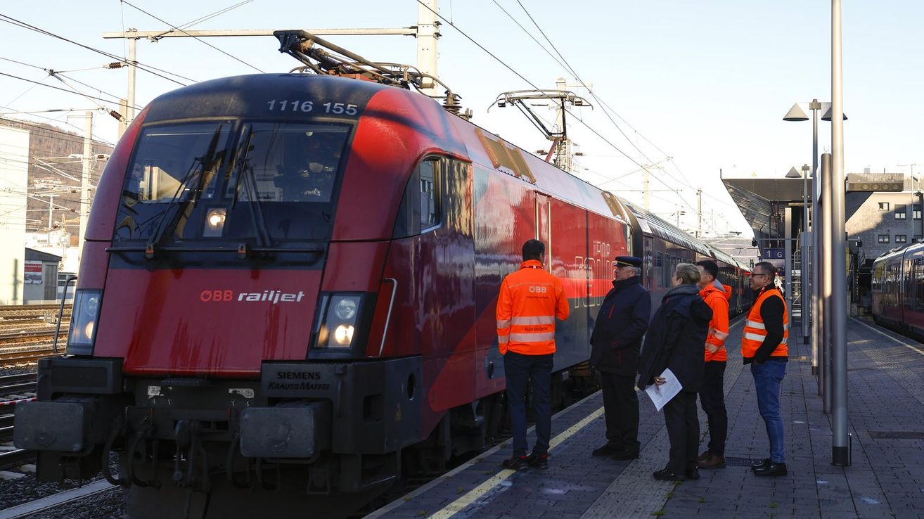 Ein roter Zug mit der Nummer 1116 155 steht am Bahnsteig, und mehrere Personen in orange Westen stehen in der Nähe. Der Zug hat das Wort railjet an der Seite.