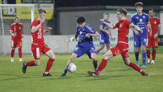 Three soccer players in action on a field, one in blue and two in red, contesting a soccer ball.