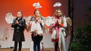 Four young girls dressed as angels with paper wings are performing on a stage. They have microphones in their ears and are standing in front of a bright red backdrop.