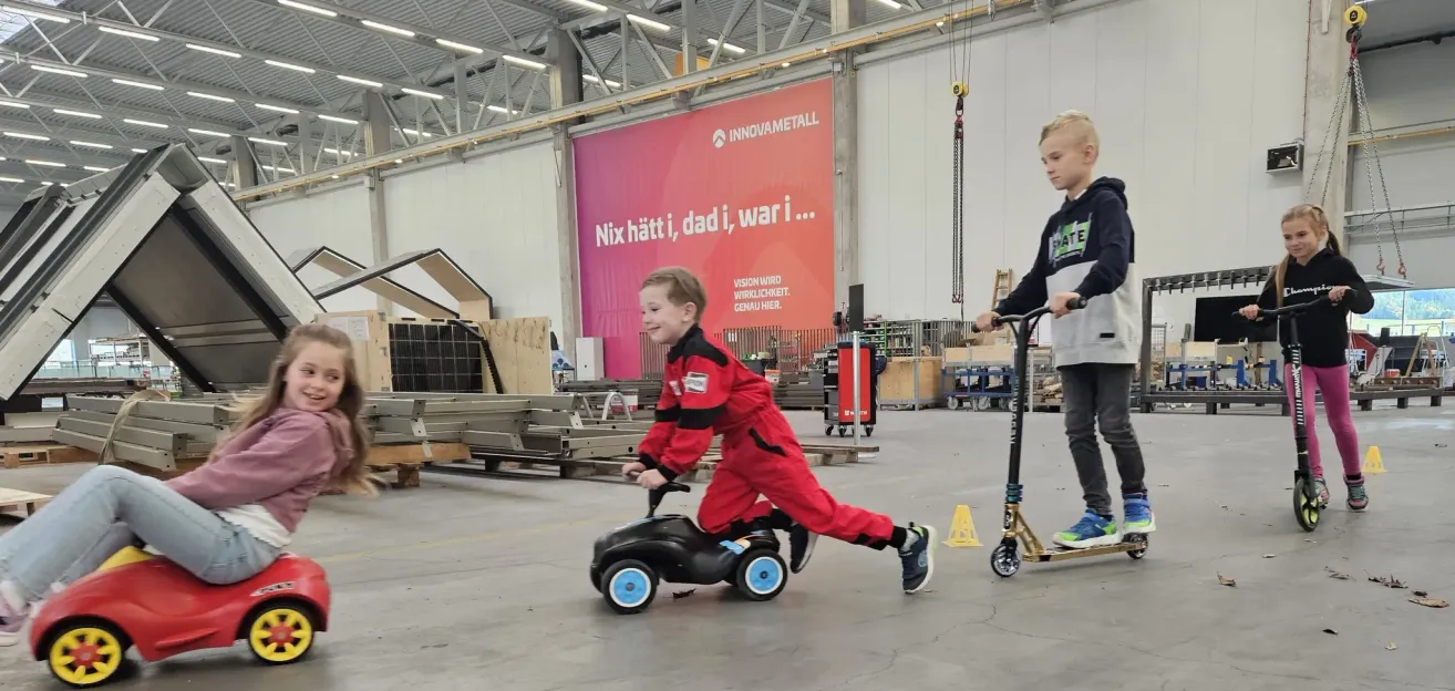 Zwei Jungen fahren auf kleinen Fahrzeugen in einem Lagerhaus, einer in einem roten Anzug und der andere in einer grauen Kapuzenjacke. Im Hintergrund befindet sich ein großes rotes Schild.