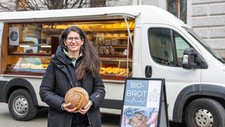 A woman with glasses and a smile holds a loaf of bread in front of a bakery van with open shelves displaying various baked goods.