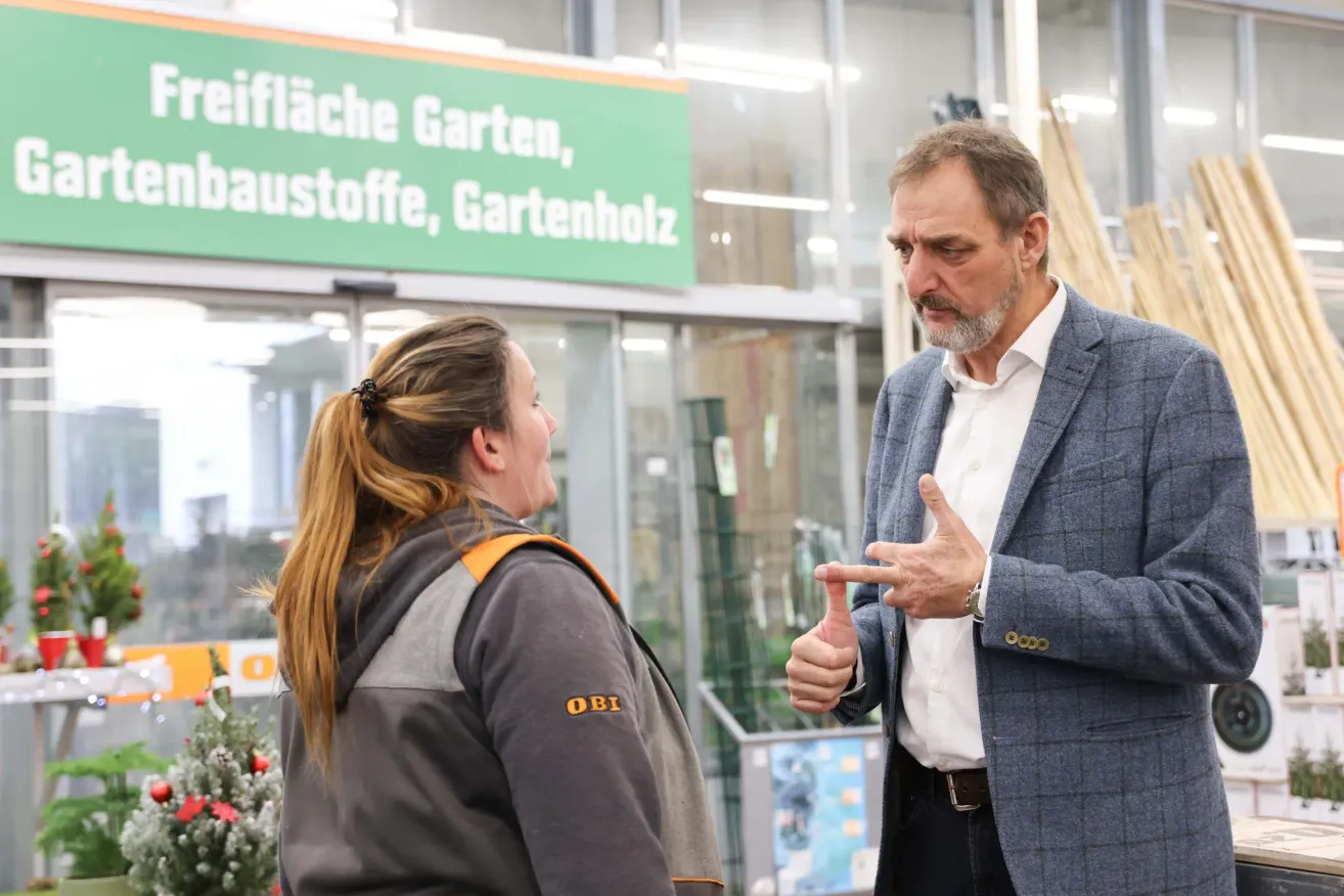Ein Mann im Anzug spricht mit einer Frau in einer Jacke vor einem Geschäft mit einem grünen Schild darüber. Das Schild hat weißen Text.