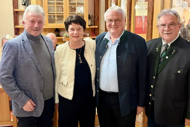 Vier Personen posieren für ein Foto, zwei Männer und zwei Frauen. Die Frau links trägt eine Halskette und eine helle Jacke. Der Mann rechts trägt ein blaues Hemd und eine dunkle Jacke. Sie alle lächeln.