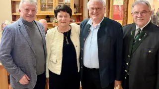 Four individuals are posing for a photograph, with two men and two women. The woman on the left is wearing a necklace and a light-colored jacket. The man on the right is wearing a blue shirt and a dark jacket. They are all smiling.