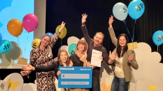 A group of adults stands on a stage with a large check and balloons. A woman holds a certificate. They are smiling and appear to be celebrating.