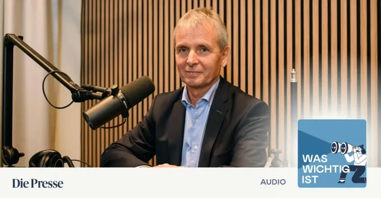 Ein Mann in einem dunklen Anzug sitzt vor einem Mikrofon, wahrscheinlich für ein Podcast oder Interview. Er hat kurzes, graues Haar und ein blaues Hemd. Das Mikrofon steht auf einem Ständer hinter ihm. Der Hintergrund ist eine Holzwand.