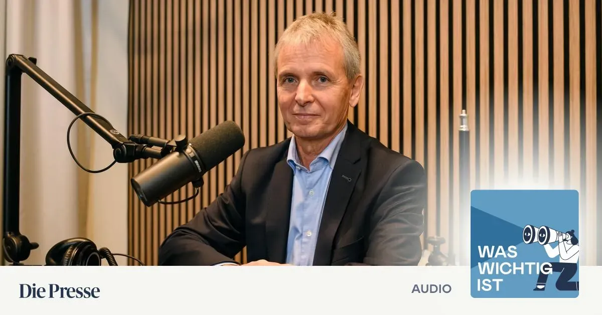 Ein Mann in einem dunklen Anzug sitzt vor einem Mikrofon, wahrscheinlich für ein Podcast oder Interview. Er hat kurzes, graues Haar und ein blaues Hemd. Das Mikrofon steht auf einem Ständer hinter ihm. Der Hintergrund ist eine Holzwand.