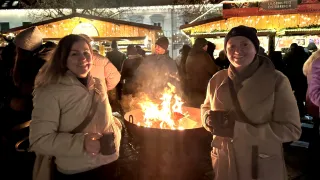 Two women stand by a fire at a Christmas market, smiling. Other people, some in hats, stand nearby, and festive lights adorn the background.
